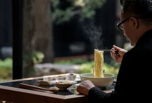 An Asian man sits at a wooden dining table with a bowl of noodles, surrounded by cups, spoons, and vegetables in a warm and inviting atmosphere