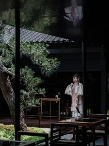 A woman in traditional Chinese clothing stands in front of a table with a vase, surrounded by chairs in a restaurant setting, conveying cultural heritage and hospitality