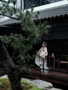 A woman in traditional attire stands in front of a building with a wooden table and chairs, surrounded by potted plants, conveying a sense of tradition and cultural significance