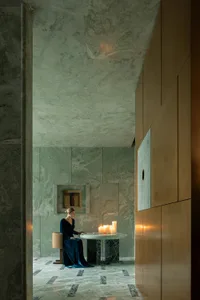 A woman reads a book by candlelight in a dimly lit room with a stone wall and a chair