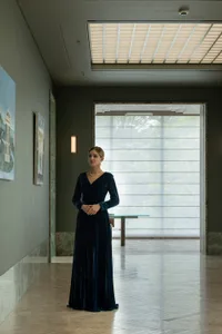 A woman in a long dress poses in front of a painting in an art gallery with a green wall, a window, chairs, and a dining table