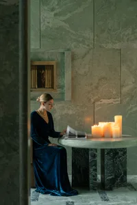A woman sits at a table in a dimly lit room with a stone wall, surrounded by candles and a vase, with two chairs and a book