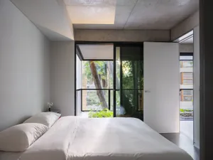 A modern bedroom with a white bed in front of a window, surrounded by concrete walls, potted plants, and a chair, creating a calm and inviting atmosphere