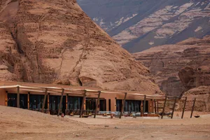 A restaurant or hotel with a large outdoor dining area featuring multiple tables and chairs on a sandy surface, surrounded by potted plants and a mountain range in the background