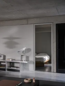 Modern minimalist bedroom with concrete walls and floors, featuring two beds, a chair, and a dining table in a clean and organized space