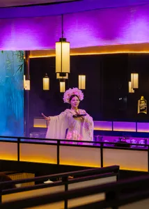 A woman in traditional attire stands on a stage in a purple-themed restaurant, surrounded by bowls and bottles, holding a plate of food and wearing a flower-adorned hat