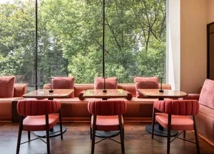 Dining area with multiple tables, chairs, and a bench, set against a backdrop of wooden floors and warm brown and white walls, bathed in natural light