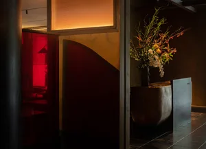 A warm and inviting interior space with a red accent wall, a large vase with flowers, and three chairs arranged around the area, surrounded by potted plants and greenery