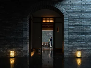 A woman stands in front of a doorway in a traditional Asian restaurant with stone walls, a brick archway, and cozy dining chairs