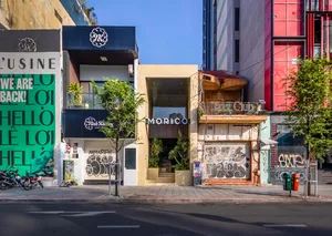熱鬧的都會街景，包含建築立面、盆栽植物與前方兩張長椅，營造溫馨氛圍