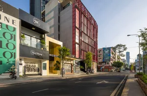 現代城市街景，擁有紅色點綴的高樓，周圍環繞盆栽植物與其他建築，背景中有兩輛機車停放在人行道上