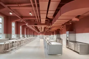 Industrial kitchen area with a large dining table and multiple chairs, surrounded by refrigerators in a commercial or industrial space