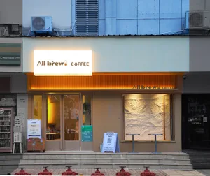 A modern coffee shop entrance with two dining tables, a bench, and scattered chairs, surrounded by white and brown walls and a potted plant