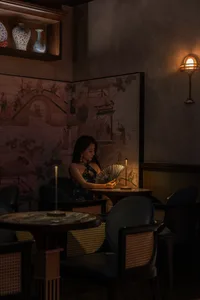 A woman sits at a dining table in an Asian restaurant with oriental artwork on the wall, surrounded by chairs and a comfortable atmosphere