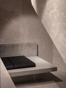 Small dark bedroom with a black bed on a concrete slab, surrounded by concrete walls and flooring, with no windows