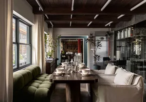 A warm and inviting living area with a dining table, couch, and decorative elements, featuring wood floors and white walls, with wine glasses on the table suggesting a social gathering