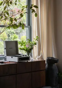 Cozy living area with a wooden dresser, record player, and natural light from a large window, featuring potted plants and a dog statue