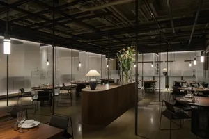 A modern restaurant dining area with wood flooring, multiple tables, chairs, and potted plants, lit by overhead lights and table lamps, creating a warm and inviting atmosphere