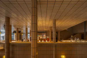 A bar area with wooden design, bottles, cups, and wine glasses on a countertop, set in a restaurant or bar with a warm atmosphere