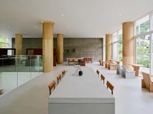 Modern dining space with concrete walls, high ceilings, and a dining table surrounded by tall columns and potted plants