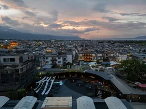 A cityscape at dusk featuring a large building with a mix of residential and commercial spaces, stone and glass facade, and a vibrant urban atmosphere