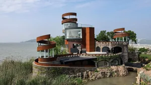 A modern house on stilts over water features red brick and metal elements, stone walls, and a serene waterfront setting