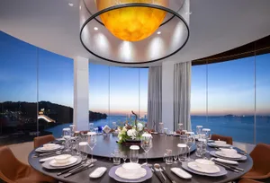 A dining area with a round table, chairs, wine glasses, plates, and cutlery, set against a warm background with a couch and potted plant, suggesting a sunset atmosphere