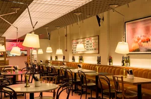 A restaurant interior with multiple dining tables and chairs, inviting atmosphere, and bottles on tables, with a clock on the wall
