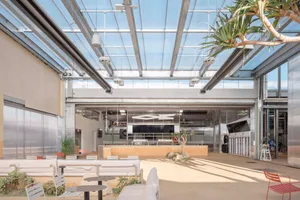 A modern open space with high ceilings, a kitchen area, and a dining area, featuring well-lit modern design, chairs, a couch, and a potted plant