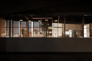 Industrial kitchen with two ovens, three refrigerators, and two people working, suggesting a busy and well-equipped commercial space