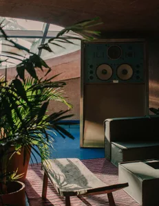 Comfortable living area with a couch, chair, and refrigerator, surrounded by potted plants and natural light from windows