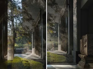 A house with a calm atmosphere, featuring large windows with blinds and a smaller window with curtains, and a bench near the windows, surrounded by trees and sunlight