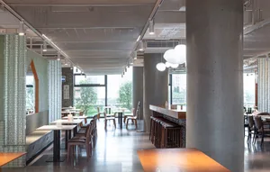 A modern restaurant interior with a large dining area, concrete elements, wood accents, and potted plants, creating a warm and welcoming atmosphere