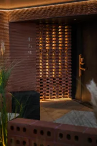 A traditional Asian-style doorway with wooden panels and a metal door handle leads to a dimly lit room with a brick wall, surrounded by potted plants