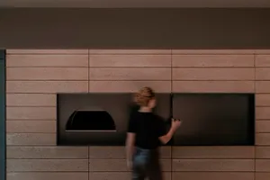 A woman walks past a brick wall in an industrial kitchen with two ovens and a pizza on the counter