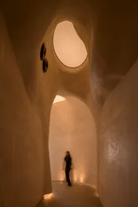A woman walks down a well-lit, curved concrete hallway with two clocks on the wall, one near the left side and one further back