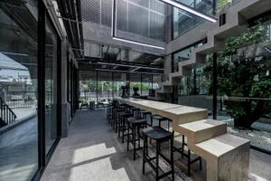 A modern bar area with a long counter and stools, surrounded by chairs and featuring concrete as a prominent material, with warm lighting creating a welcoming atmosphere