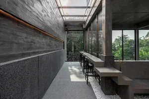 A modern restaurant with a large dining table, concrete walls, a glass ceiling, and potted plants, creating an inviting atmosphere for socializing and dining