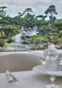 A large white building with a spacious patio area, dining table, and potted plants, surrounded by a waterfall and a bird in flight