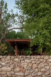 A rustic house with a stone wall in front, surrounded by trees, featuring a wooden structure and brick exterior