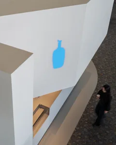 A modern interior space with a white wall featuring a blue glass bottle shape, two people, and a cup, set in a minimalist atmosphere