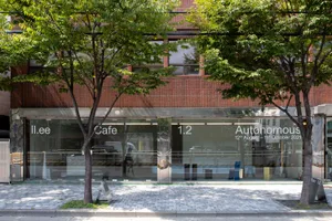 A modern cafe building with a brick exterior, a large window reading 'Cafe', and several cars parked outside, surrounded by trees and a city atmosphere