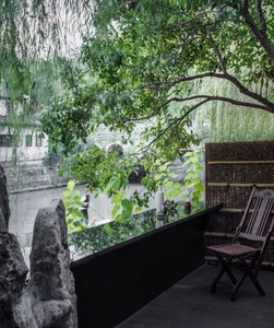 A serene outdoor patio area with a wooden chair on a stone surface near a tree, surrounded by other chairs and lush greenery