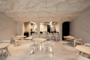 Open modern kitchen and dining area with white walls and floors, featuring a dining table and chairs, and a kitchen section with a refrigerator and oven