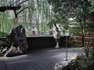 A serene garden area with a woman in a white dress standing near a large rock, surrounded by potted plants and two chairs, with a peaceful atmosphere