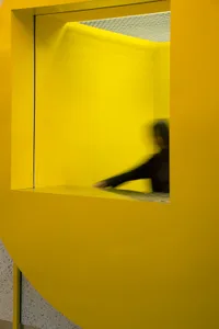 A woman stands in front of a yellow wall, possibly interacting with a door or object, in a residential or public building with a distinctive design element