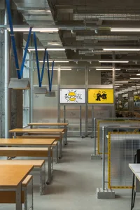A modern office space with multiple tables and chairs, featuring a mix of wood and metal elements, and yellow signs on some tables, set in a well-lit atmosphere
