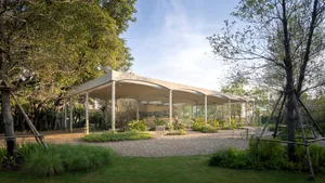 An open air living area with a large white structure featuring a glass roof, multiple rooms, chairs, and potted plants, creating a relaxed and inviting atmosphere