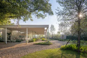 Open-air living area with a modern building featuring glass walls, potted plants, and chairs, blending indoor and outdoor spaces