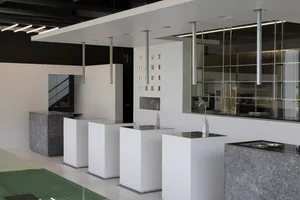 A modern bathroom with white fixtures, three sinks, two toilets, and a large glass wall, giving a clean and minimalist impression with natural light